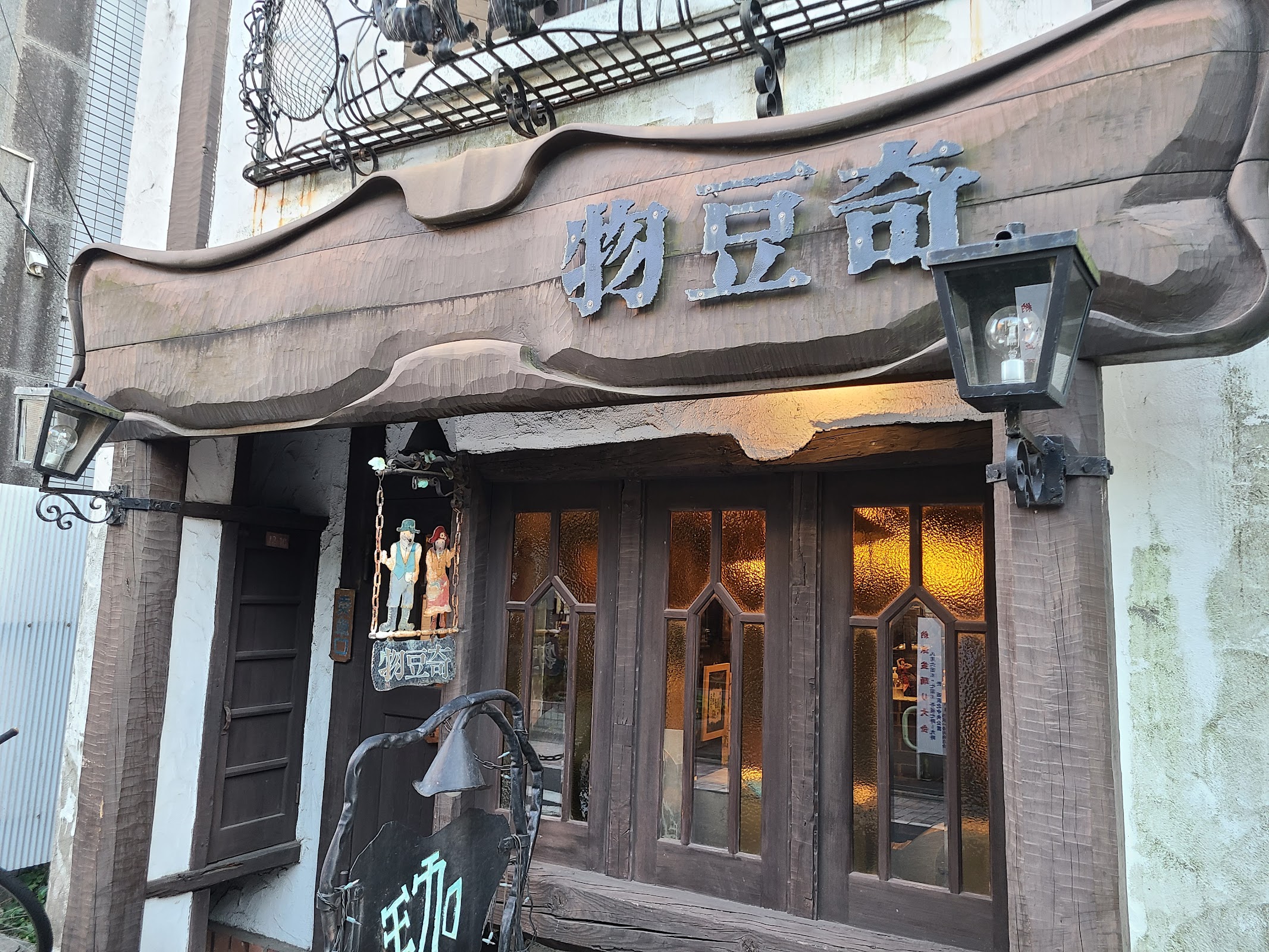 木の看板に「物豆奇」の文字、両脇のガス灯風ランプと木枠ガラス扉が琥珀色に光る店頭クローズアップ