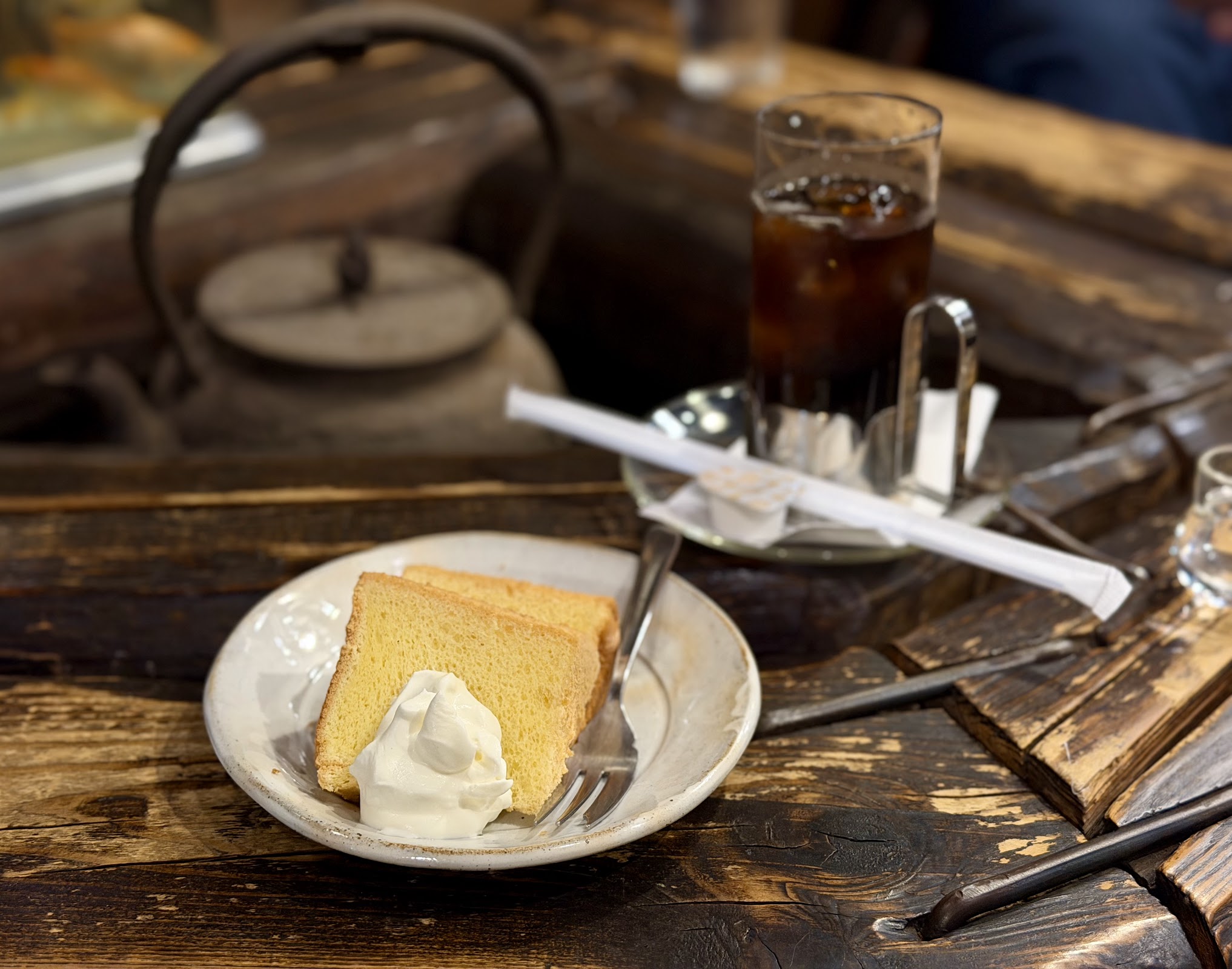 白い陶器プレートにシフォンケーキ2切れとホイップクリーム、奥にアイスコーヒーのグラスと鉄瓶が置かれた囲炉裏風テーブル